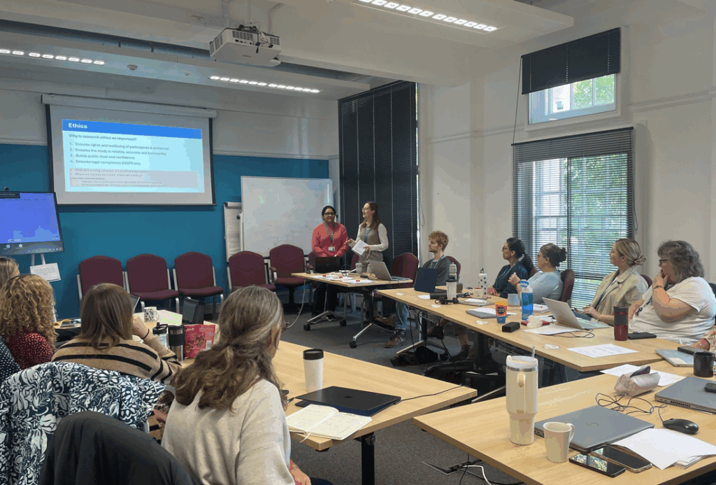 A group of people sit around tables in a classroom while two presenters stand at the front delivering a presentation displayed on a screen.