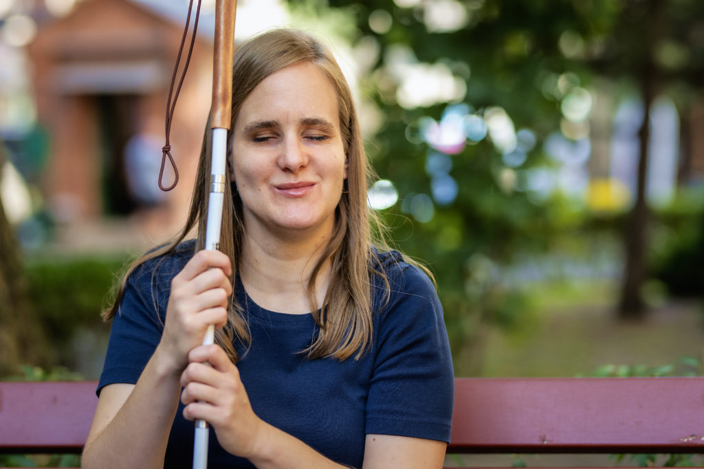 Close-up of a visually impaired woman holding a white cane and sitting on a bench in the city. Smiling female wearing casual clothes.