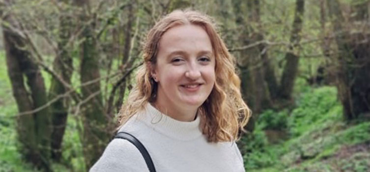 Abbit is standing in a wooded area, she has blonde curly hair and is wearing a white high neck jumper