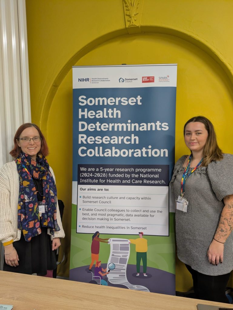 Two women stand in front of a poster promoting the Somerset Health Research Collaboration.